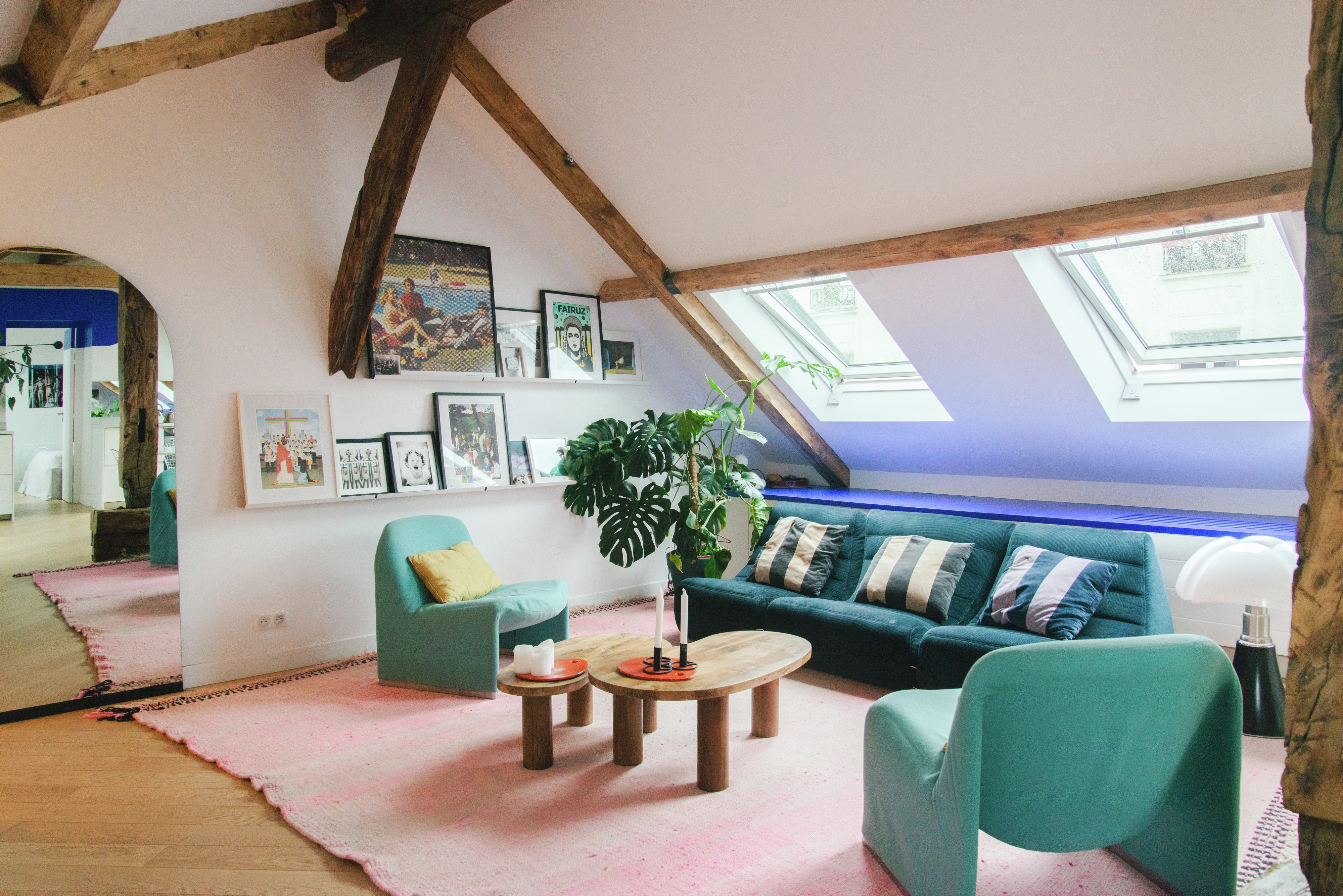 Salon de comble élégant avec des fenêtres VELUX, un canapé bleu, des poutres en bois et des œuvres d'art encadrées.