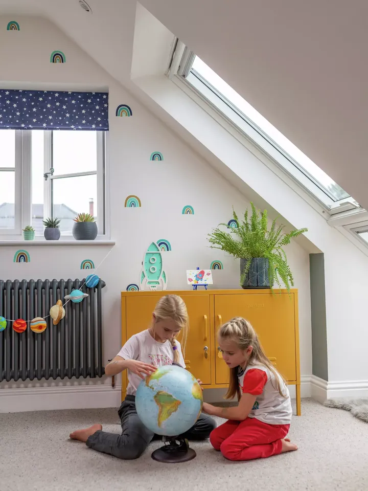 Kinder in einem hell erleuchteten Raum mit einem VELUX-Fenster, die einen Globus neben einem gelben Schrank erforschen.