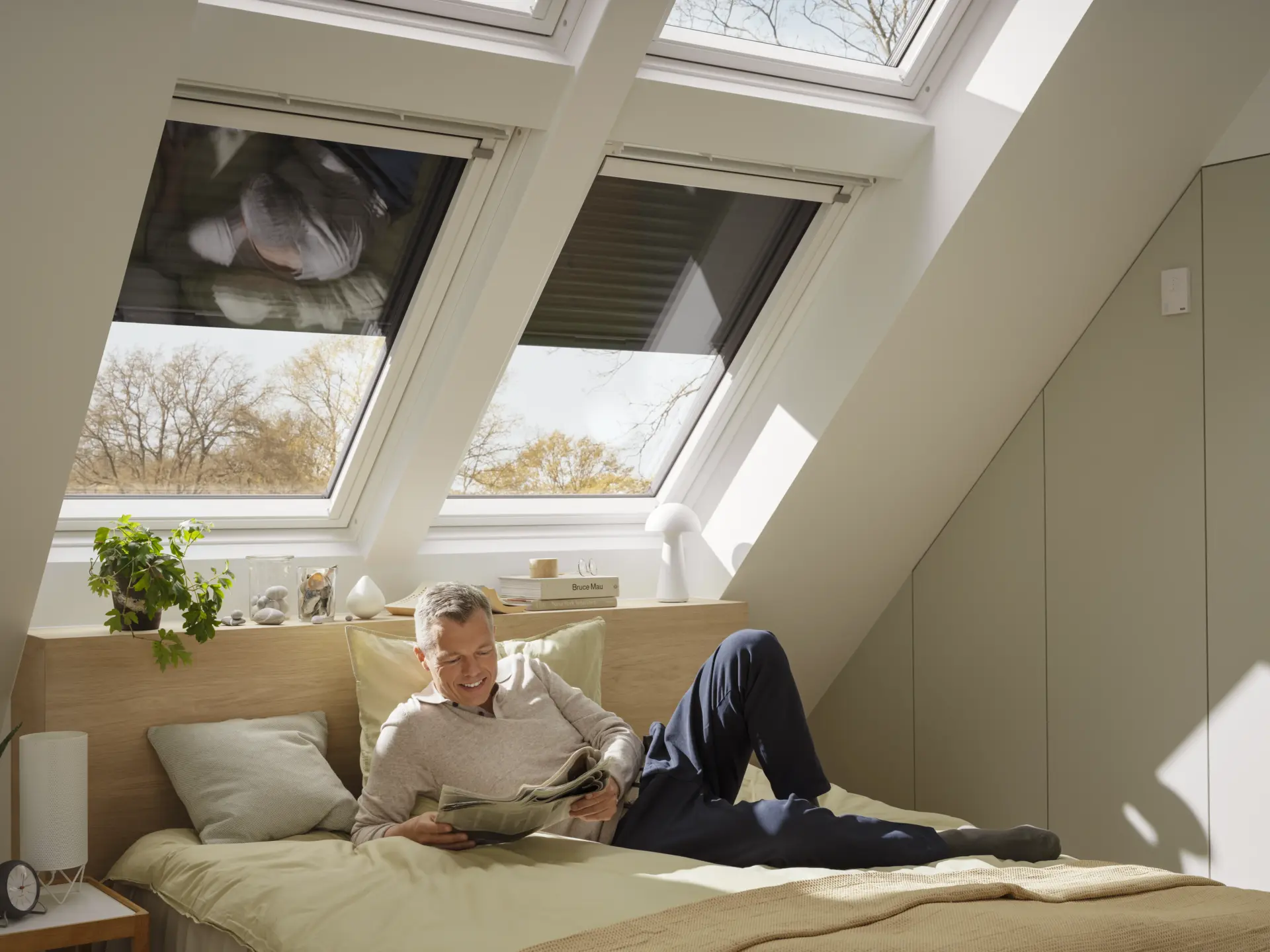Férfi olvas világos tetőtéri hálószobában, ahol a VELUX tetőtéri ablakokon külső hővédő árnyékolók segítenek csökkenteni a nyári felmelegedést és kellemes beltéri klímát biztosítanak. 