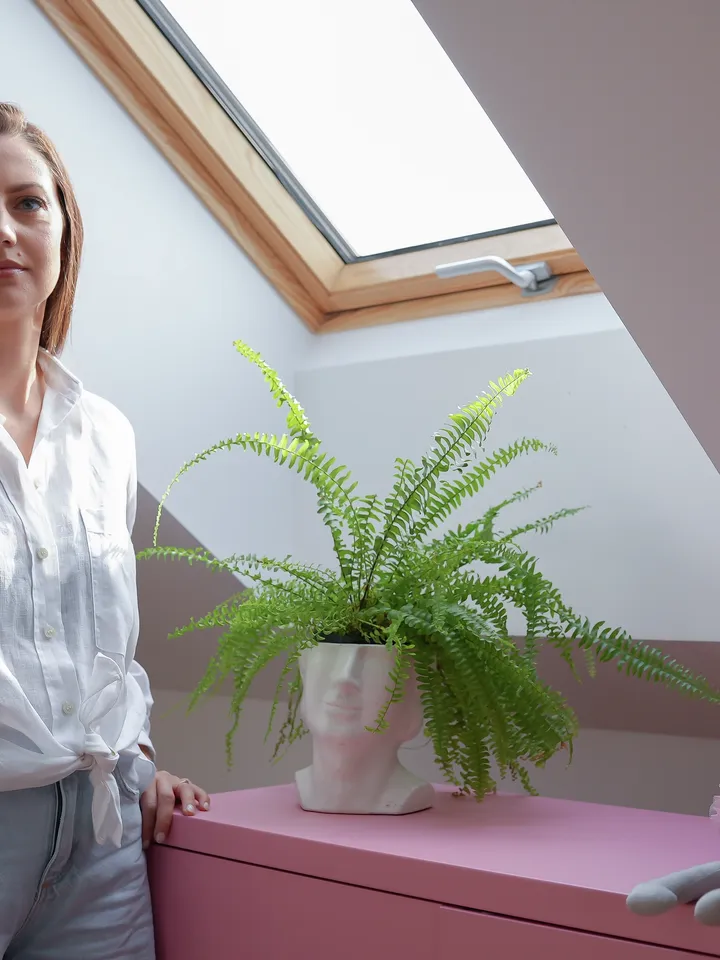 Kobieta stojąca pod oknem dachowym w jasnym pokoju na poddaszu z rośliną i pluszowym kotem | VELUX