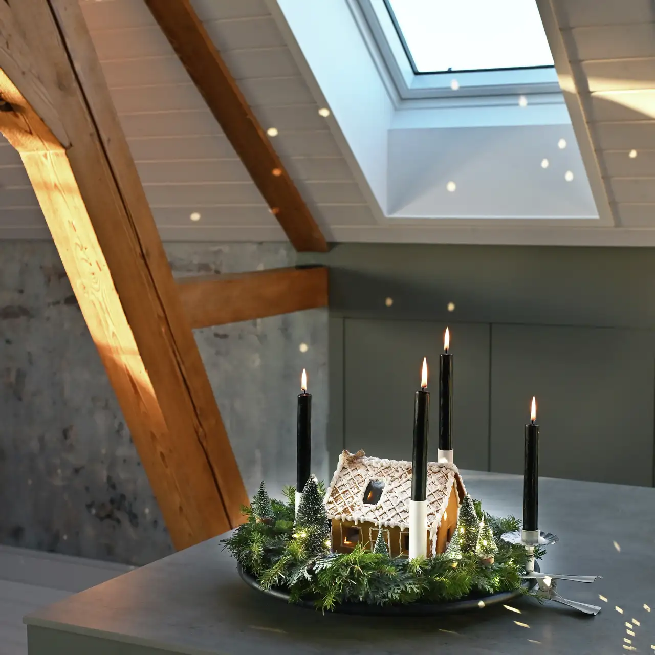 Candele nere accese in una composizione attorno a una casetta di pan di zenzero posizionata sotto una finestra per tetti VELUX
