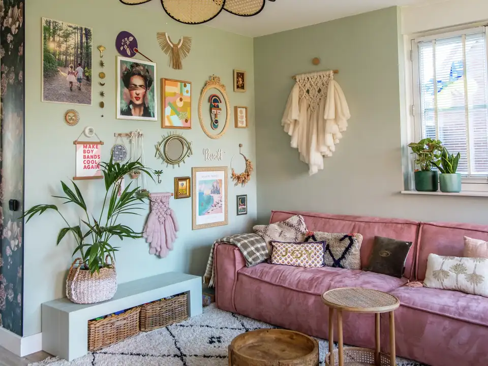 Soggiorno arredato in stile boho, con parete verde salvia, cornici, divano rosa etappeto