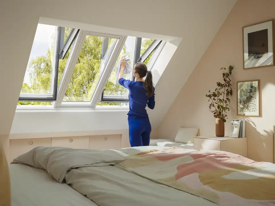 Attic bedroom with VELUX roof window open, woman cleaning the window.