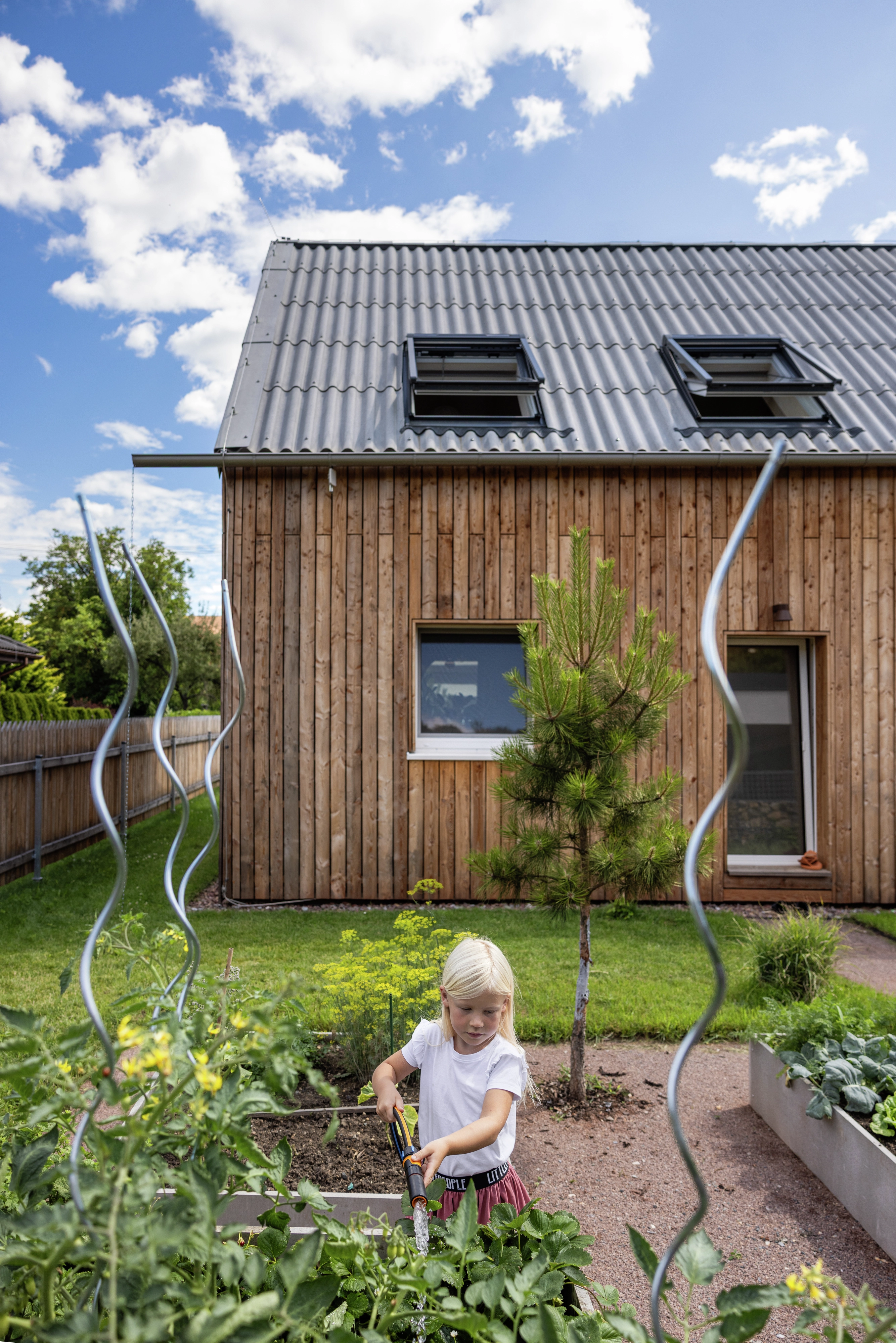 Child watering plants in garden, wooden house with metal roof and VELUX windows.