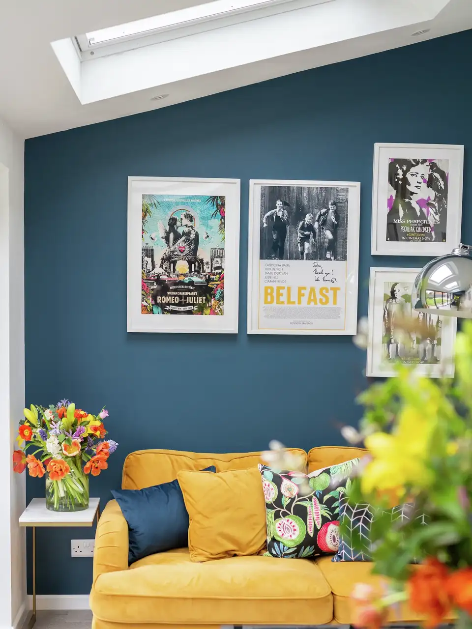 Coin salon lumineux avec fenêtres de toit VELUX, mur bleu profond, canapé jaune curry et cadres décoratifs personnalisés
