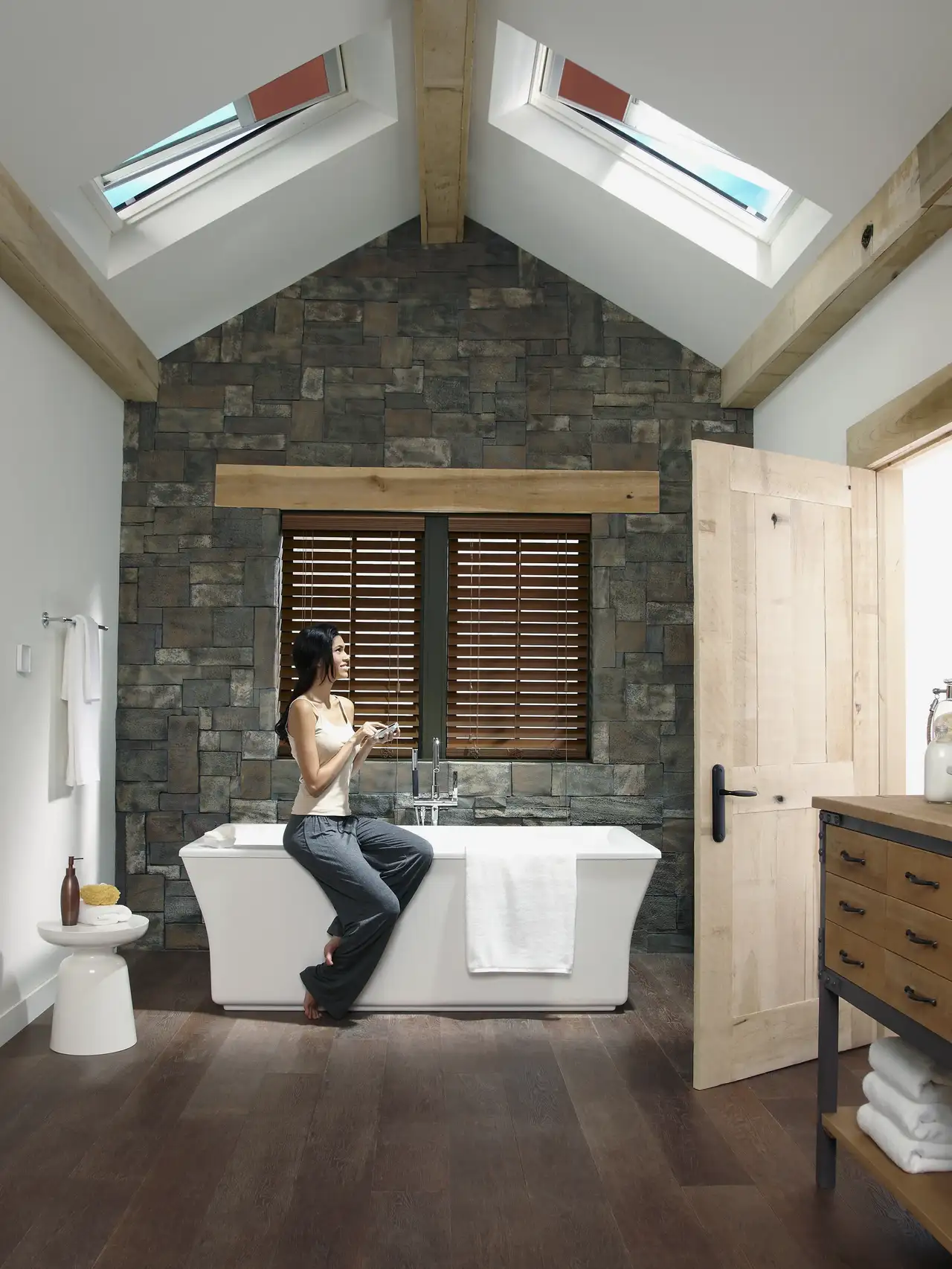 Salle de bain avec baignoire îlot et murs en pierre, éclairée par des fenêtres de toit équipées de stores intérieurs. 