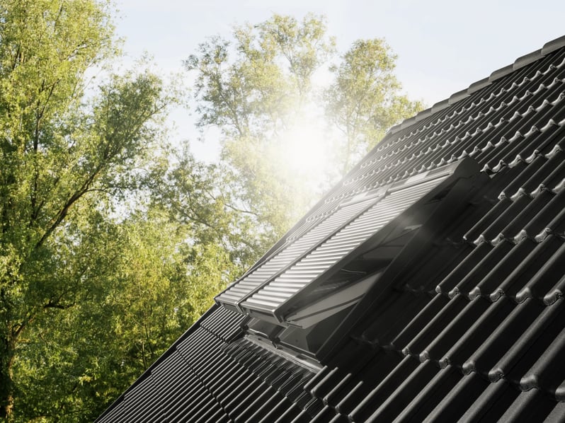 Roof with black tiles and  open VELUX roof windows with solar shuttes, surrounded by trees.