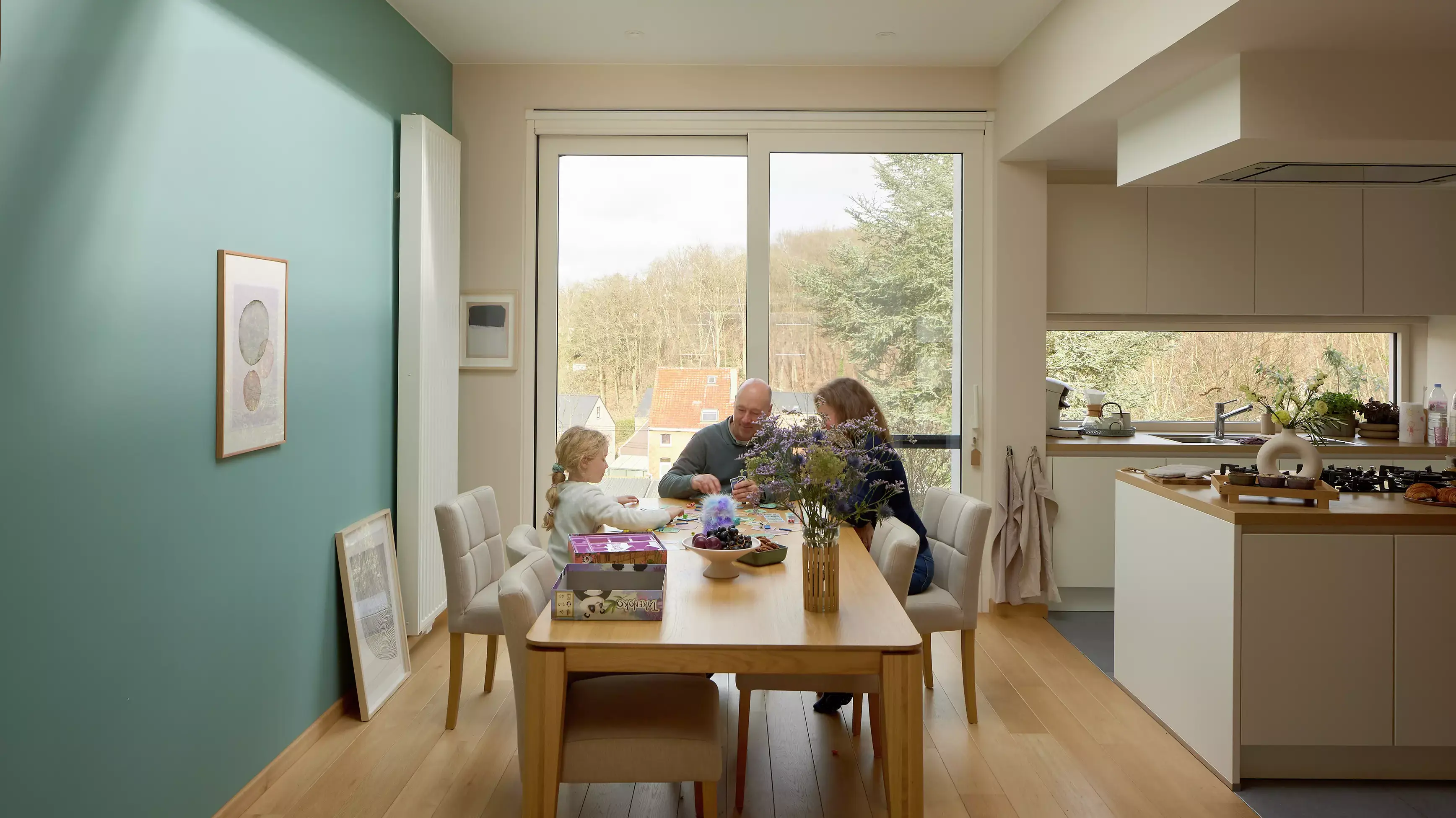Salle à manger moderne avec famille, vue sur grande fenêtre et cuisine adjacente.