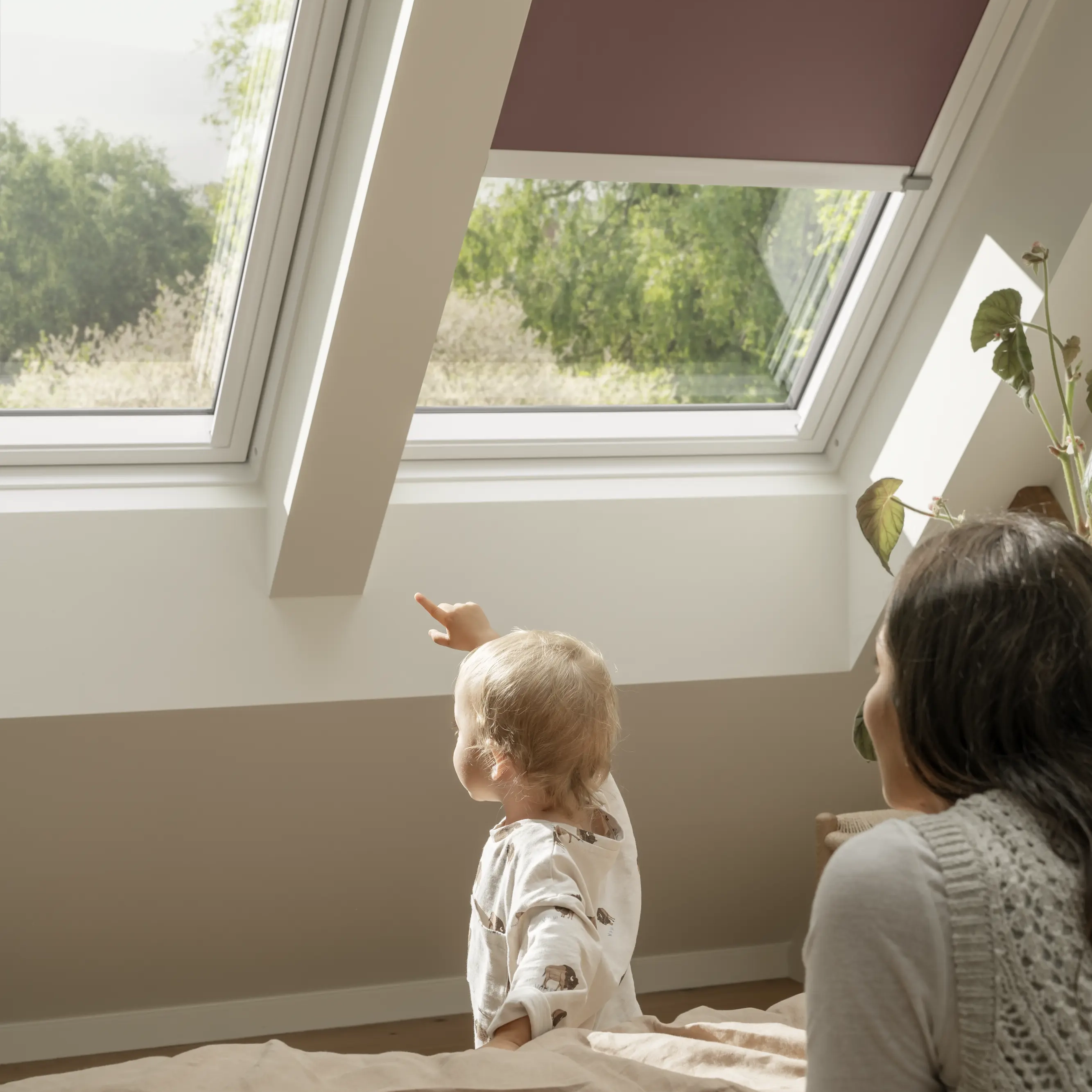 Child in bedroom pointing at VELUX roof window with greenery outside.