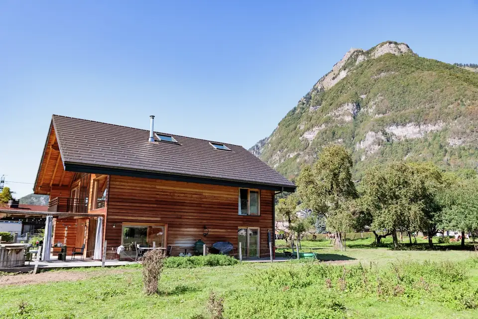 : Une maison en bois avec un toit incliné doté de fenêtres de toit, située dans un paysage verdoyant avec des montagnes en arrière-plan par une journée ensoleillée.