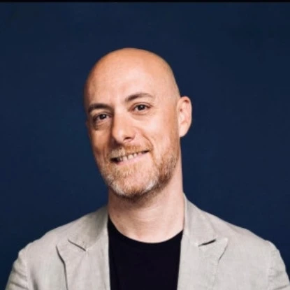 Bald man with a short beard smiling in a beige blazer against a dark blue background