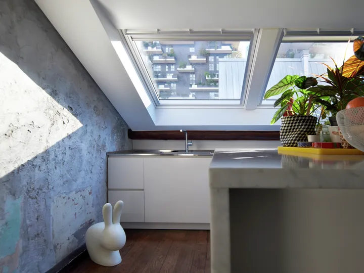 Eğimli bir çatı altında, penceler ile aydınlatılmış dokulu beton duvar, beyaz dolaplar, bitkiler ve eğlenceli bir dekoratif tavşan heykeli bulunan modern bir mutfak.