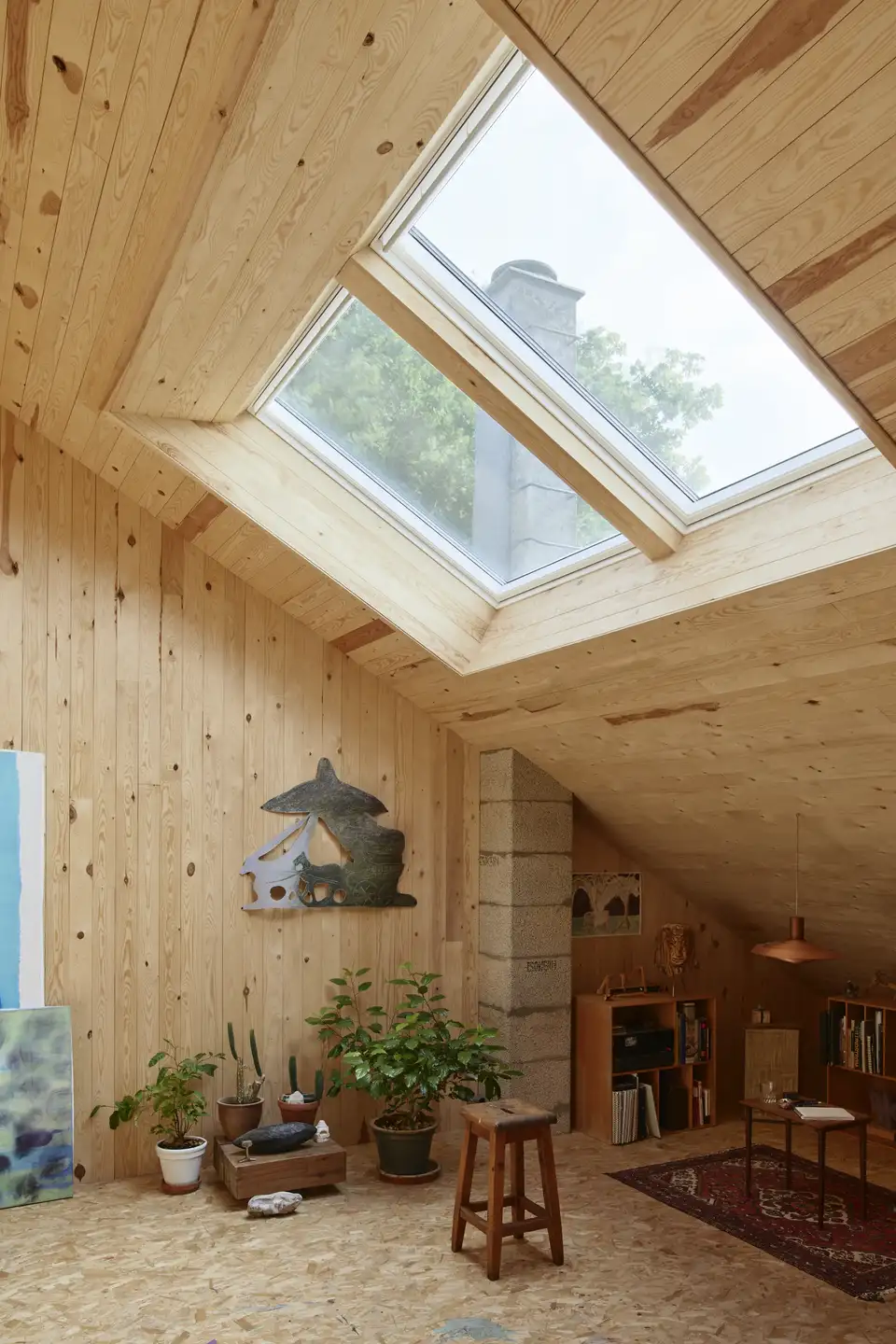 Une pièce avec un plafond incliné en bois et une fenêtre à claire-voie, des plantes éparpillées dans la pièce. Une cheminée en arrière-plan. 