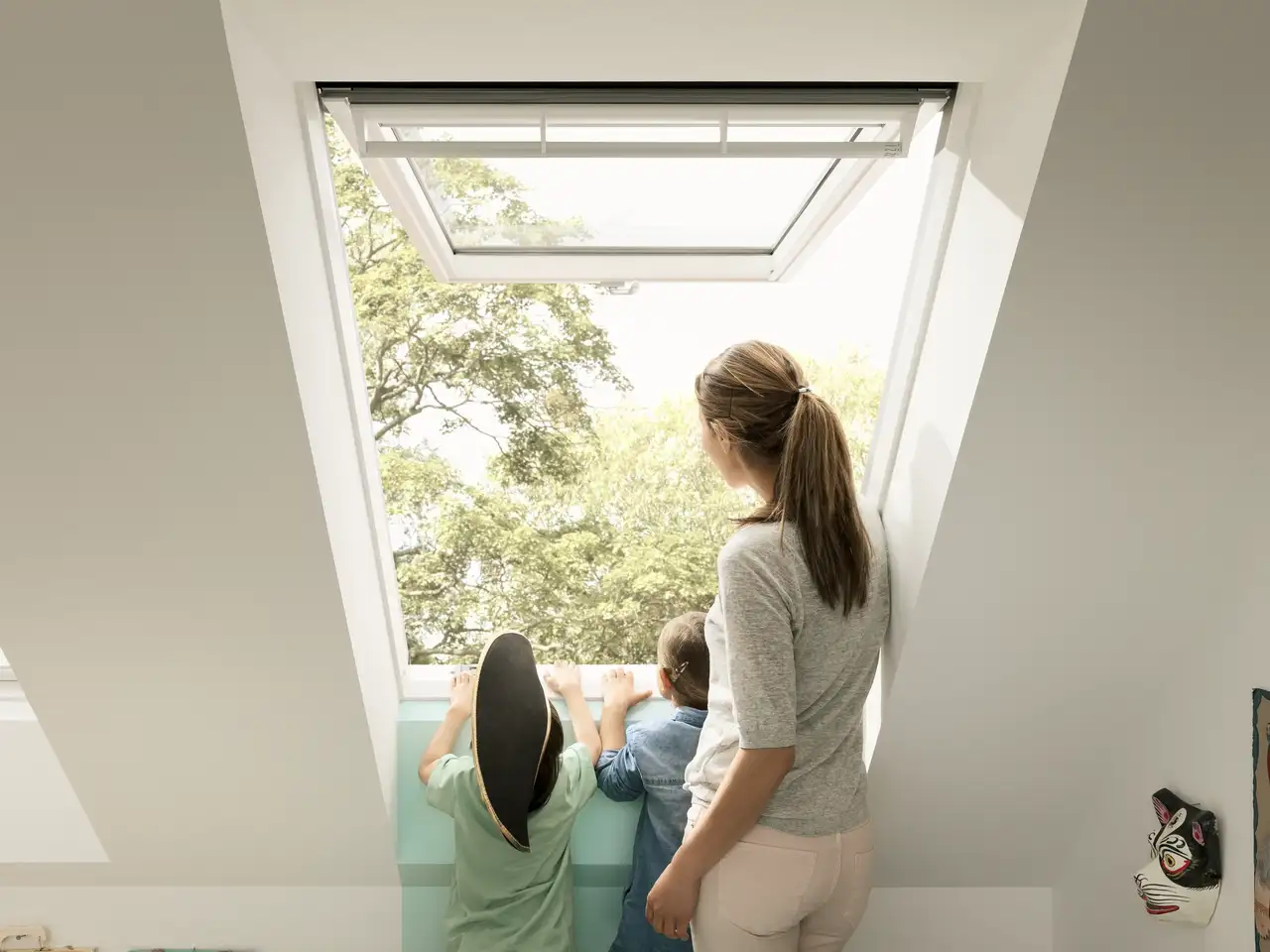 Une femme et deux enfants regardent à l’extérieur par une fenêtre de toit VELUX ouverte lors d’une journée ensoleillée 