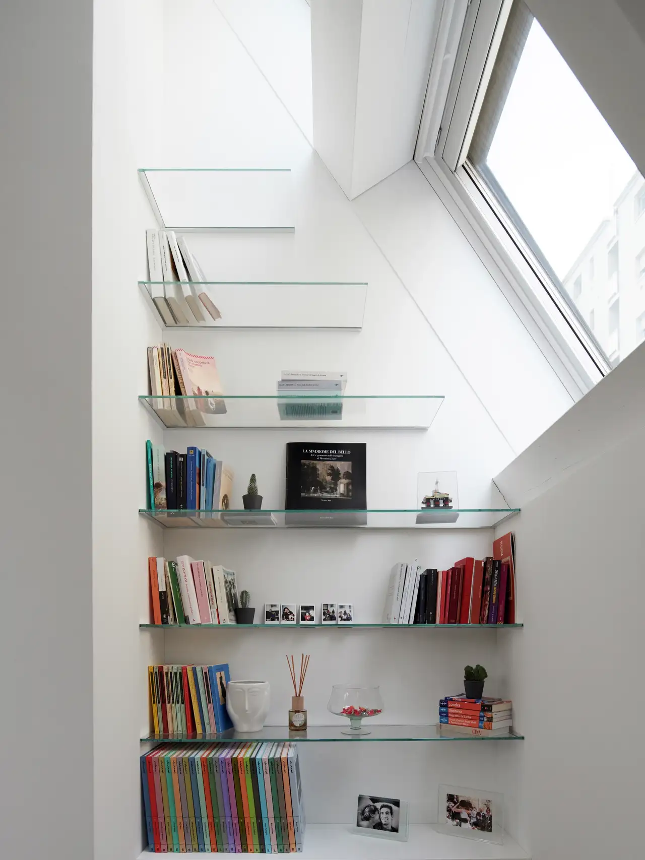 Libreria con mensole in vetro trasparente inserite in una nicchia accanto a una finestra per tetti.