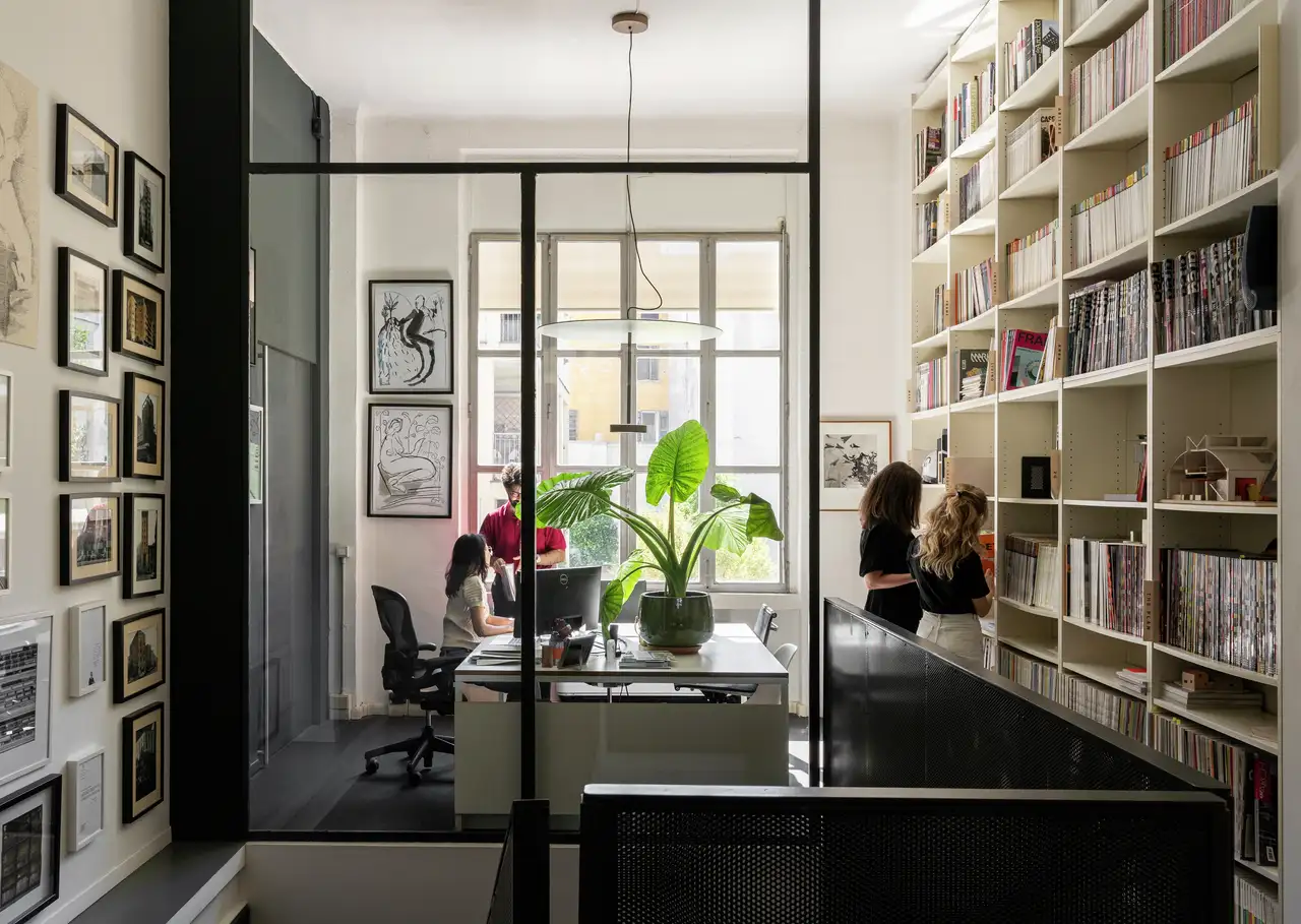 L’immagine mostra uno spazio interno, probabilmente un ufficio domestico o una sala studio. C’è una grande unità di scaffali sulla destra piena di libri e vari oggetti. Una scrivania con una sedia è posizionata di fronte all’unità di scaffali, e c’è un laptop aperto sulla scrivania. La stanza ha grandi finestre che lasciano entrare la luce naturale, e ci sono due persone visibili attraverso la parete di vetro: una seduta a un tavolo e l’altra in piedi vicino alla finestra. Lo spazio appare moderno e ben organizzato, il che potrebbe essere rilevante per scopi di design d’interni o organizzazione.