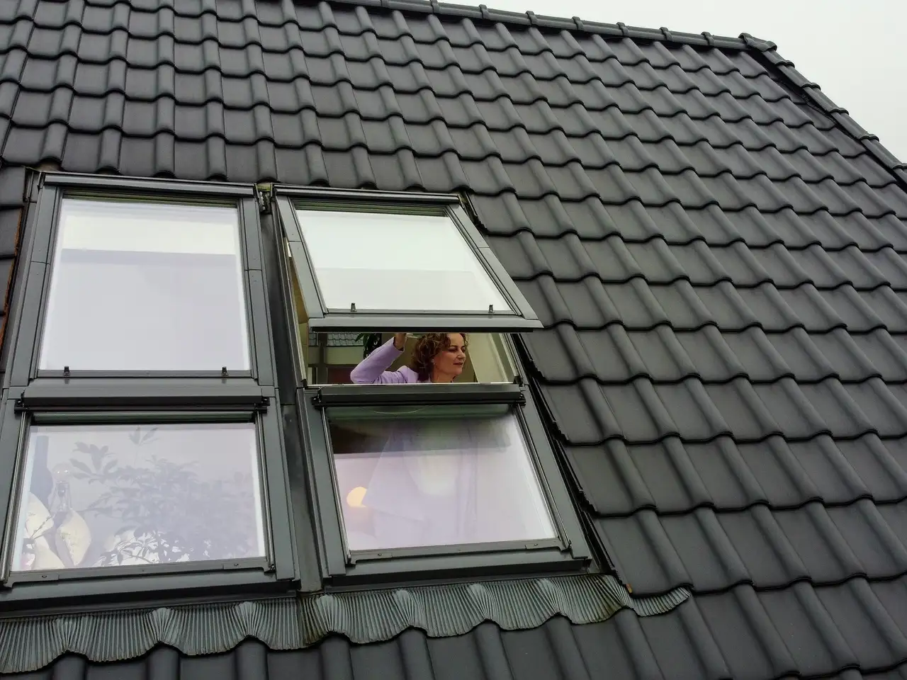 Telhado com janela de telhado VELUX aberta, telhas cerâmicas escuras visíveis. 