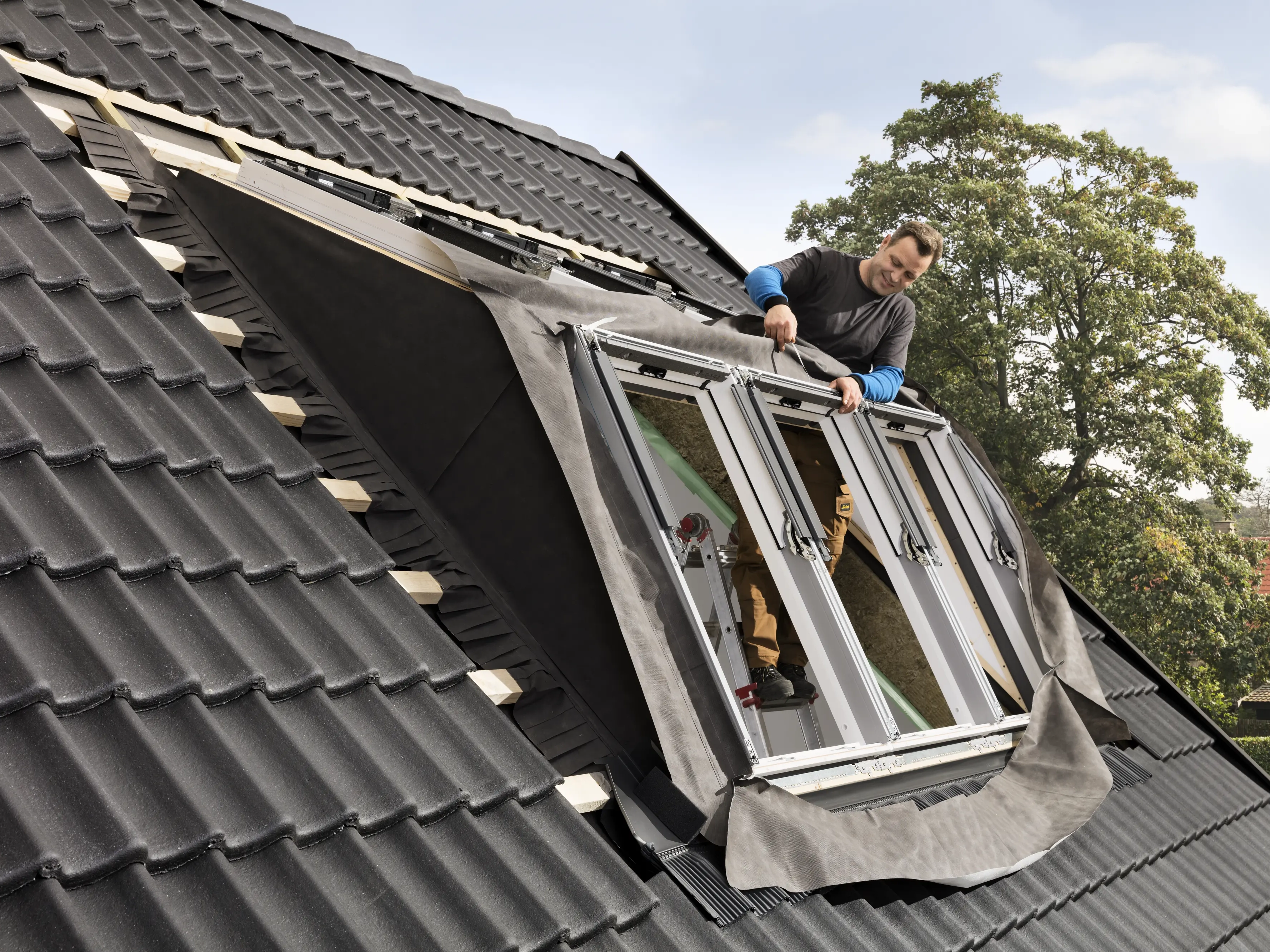 Worker installing VELUX roof window on a steep black tiled roof.