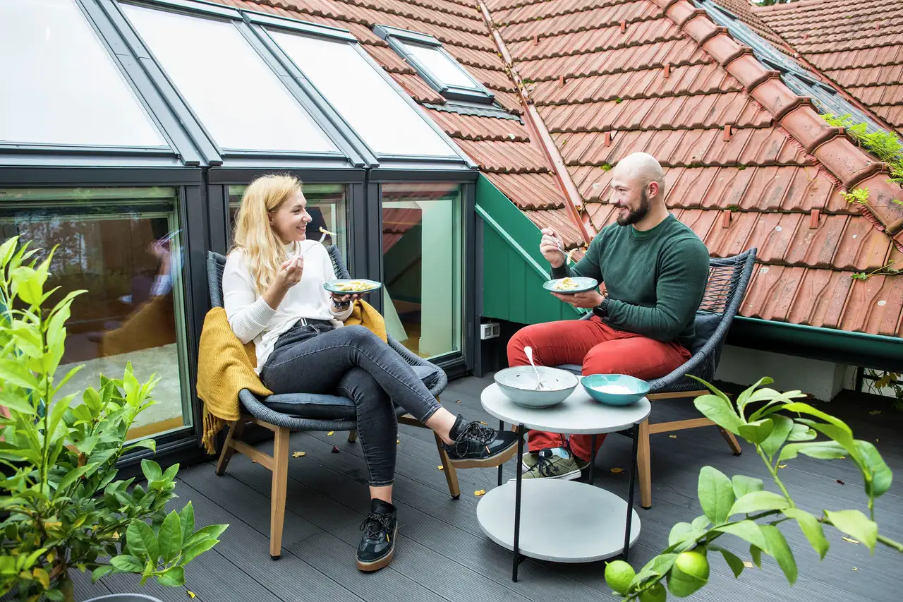 Couple enjoying a meal on a balcony with roof windows above, surrounded by greenery. | VELUX