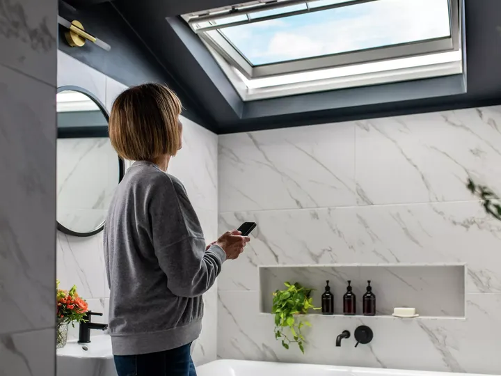 Frau in modernem Badezimmer mit Marmorwänden unter einem VELUX Dachflächenfenster, hält eine Fernbedienung.