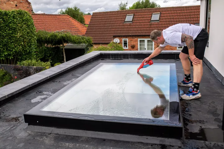 Mann mittleren Alters in sommerlicher Kleidung steht auf einem Flachdach und putzt das Flachdach-Fenster | VELUX Magazin