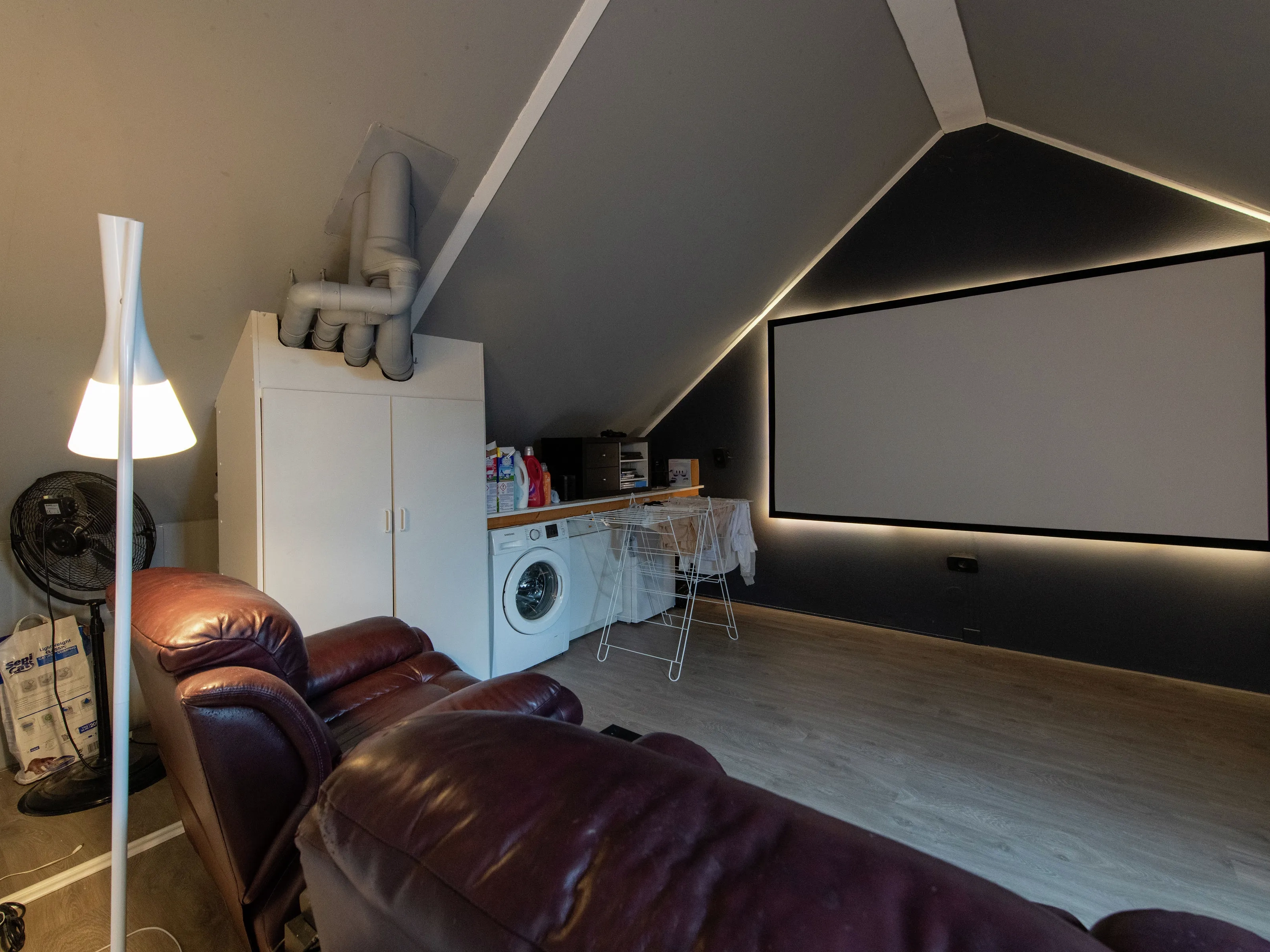 Zolder vóór de renovatie, met wasmachine en zetel onder zwakke verlichting. 