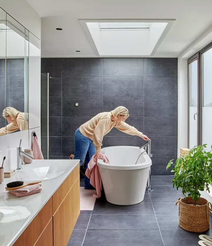 Modernes Badezimmer mit dunklen Fliesen, hölzernem Waschtisch, VELUX Dachflächenfenster und freistehender Badewanne.