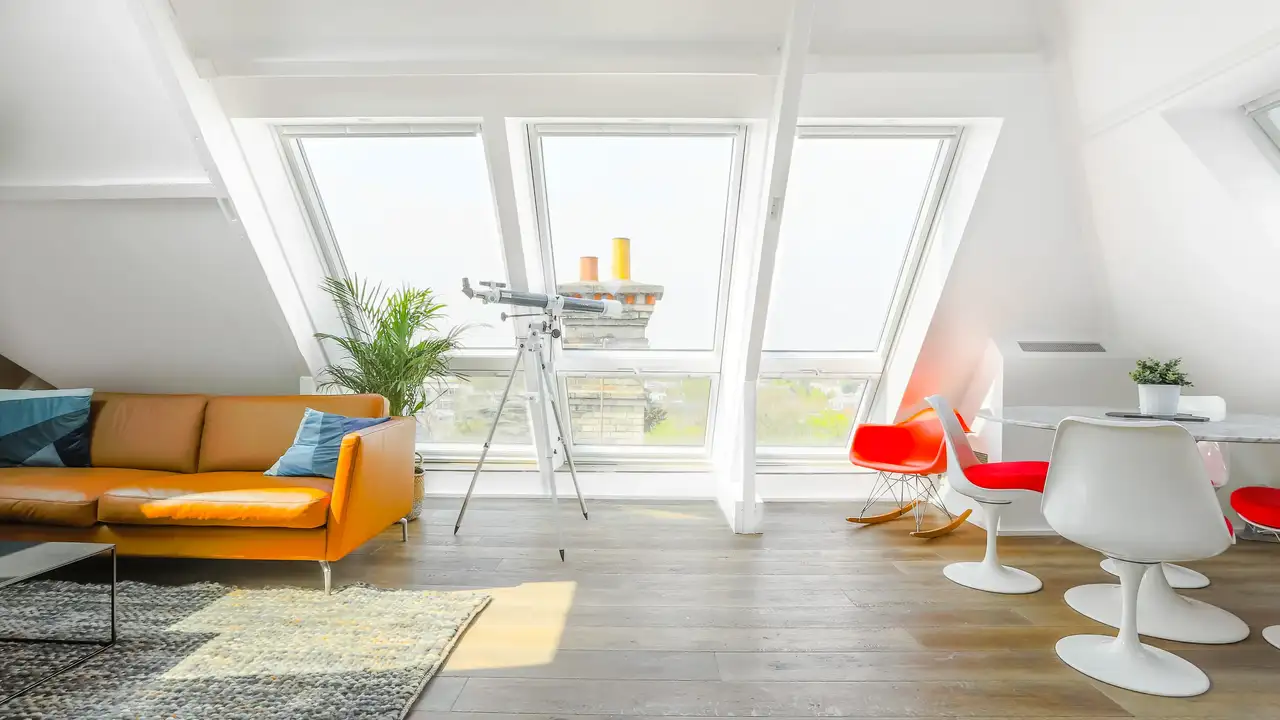 Séjour avec canapé en cuir, tapis, table et chaises rétro blanches avec coussins orange, grandes fenêtres de toit VELUX.
