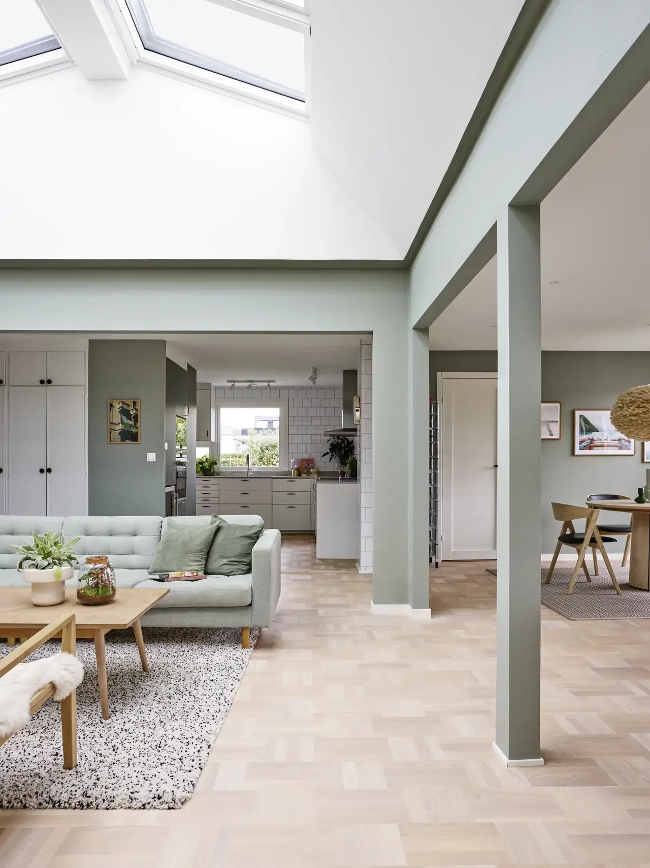 Lyst og moderne hjem med åpen planløsning, grønne vegger, lysegrå sofa og et romslig kjøkken. Dagslys flommer inn gjennom store takvinduer.