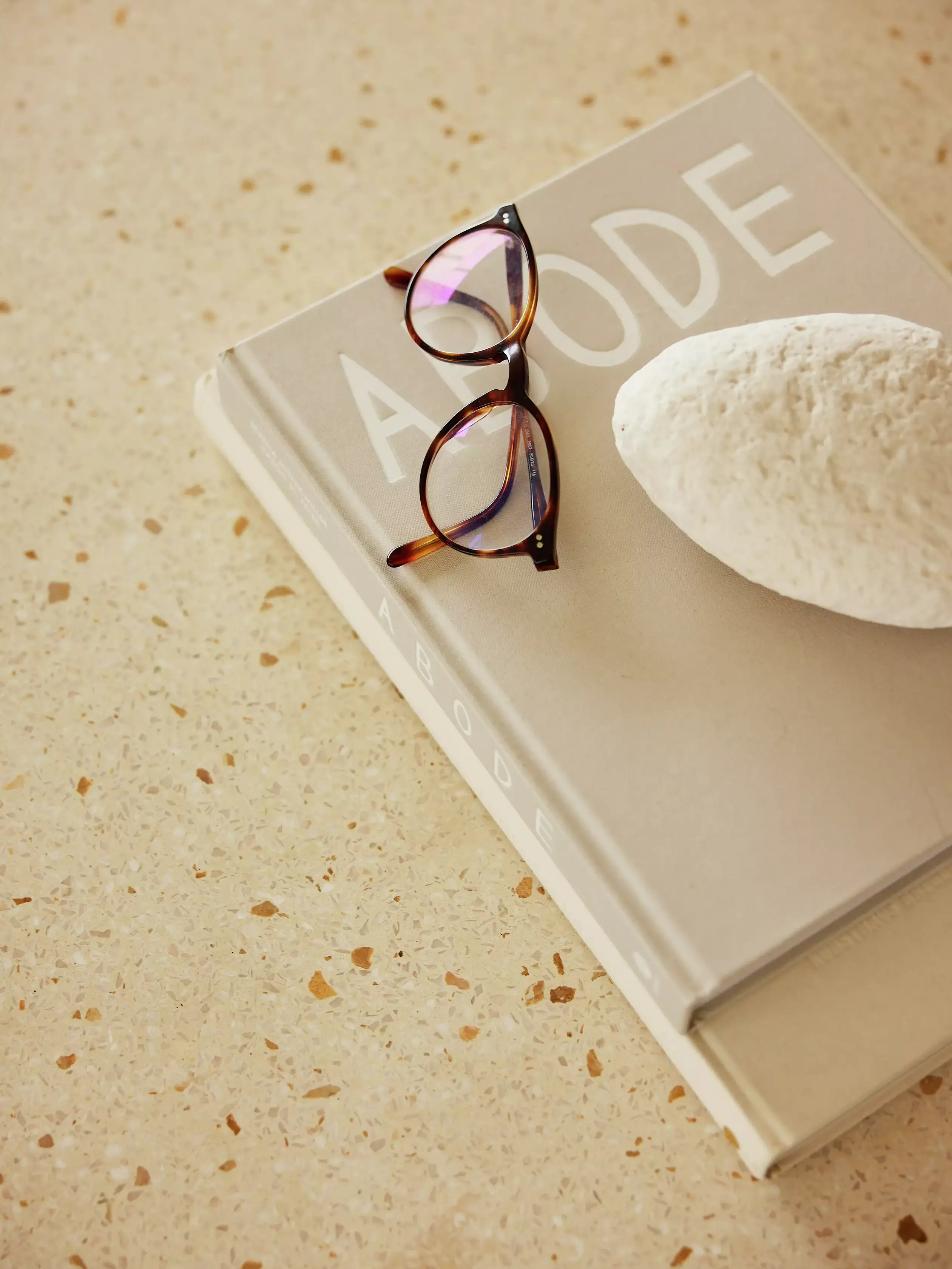 Lunettes sur le livre 'ABODE' avec un galet blanc sur une surface crème.