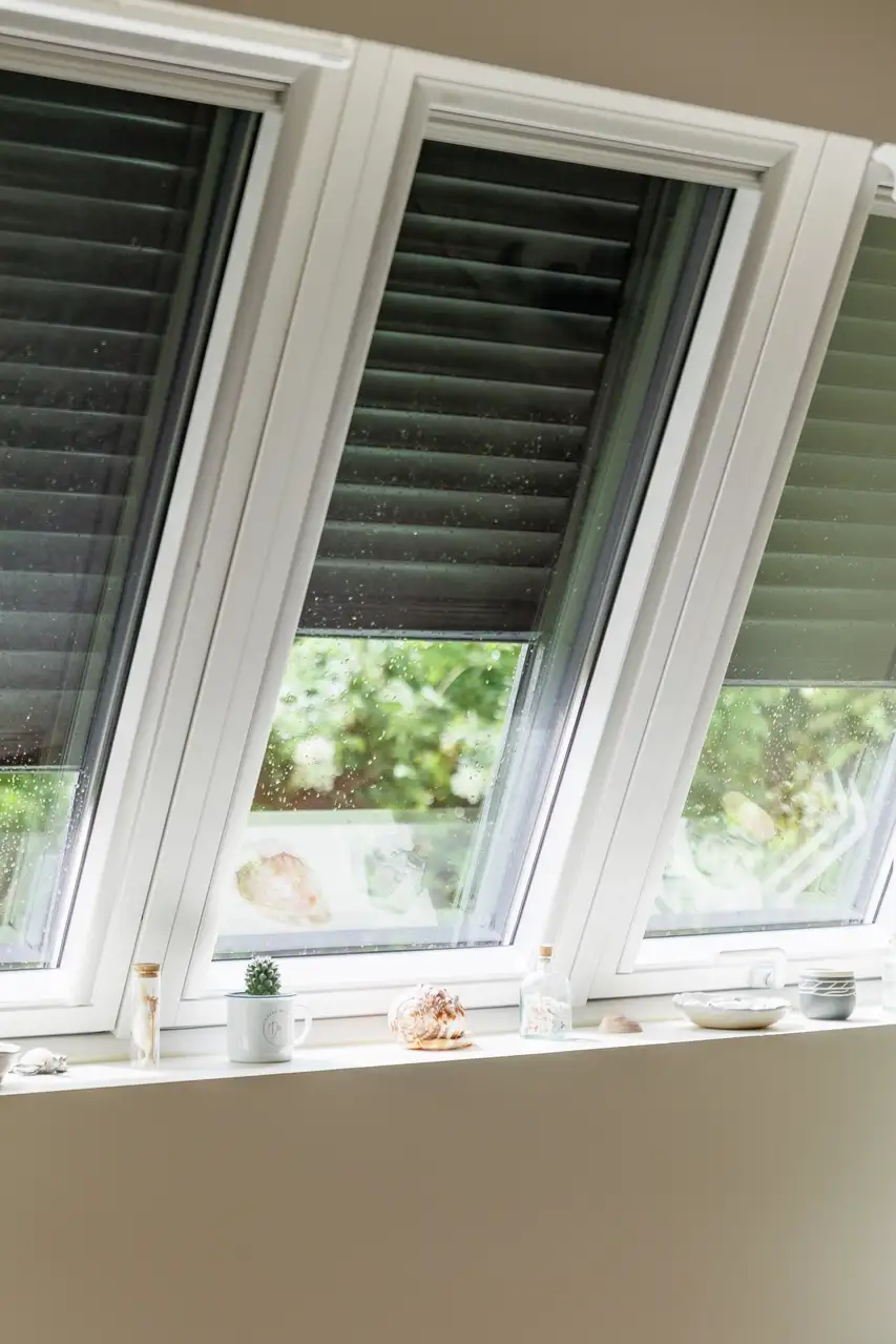Trois fenêtres de toit VELUX avec des stores partiellement fermés, un rebord de fenêtre décoré de petites plantes, coquillages et objets décoratifs. 