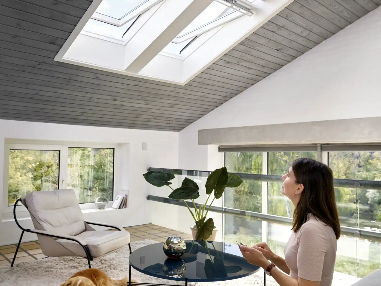 Una donna controlla nel soggiorno le Sue grandi finestre per tetti tramite l’app dello smartphone e VELUX Active. | Rivista VELUX