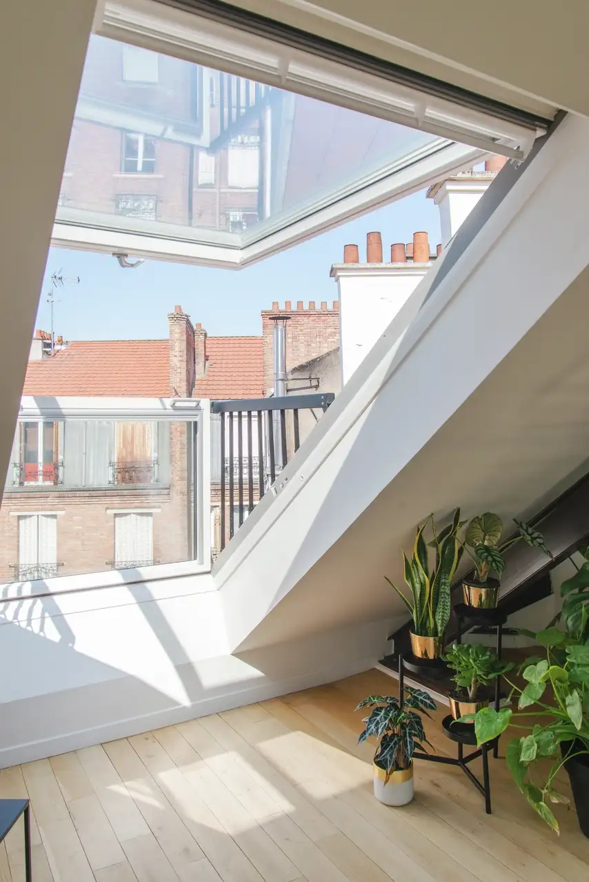 Un loft lumineux avec une grande fenêtre de toit VELUX ouverte, offrant une vue sur la skyline parisienne, entouré de plantes d'intérieur.