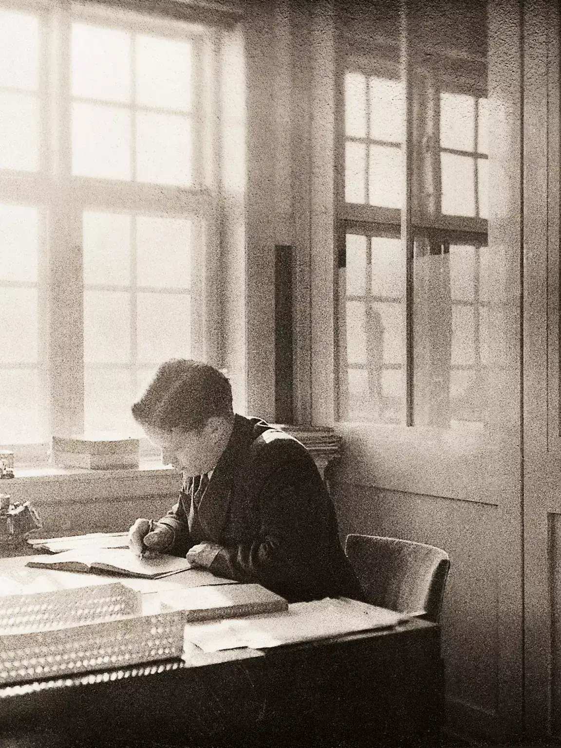 Schwarz-Weiß-Bild einer Person, die konzentriert an einem Schreibtisch nahe großer Fenster studiert.