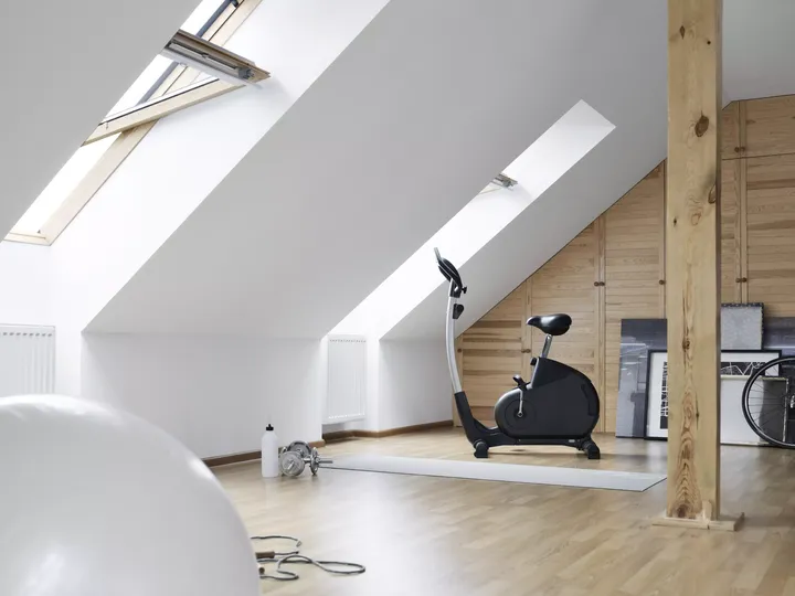 Fitnessraum im Dachboden mit einem Standfahrrad unter VELUX Dachflächenfenstern, hölzernen Balken und Boden.