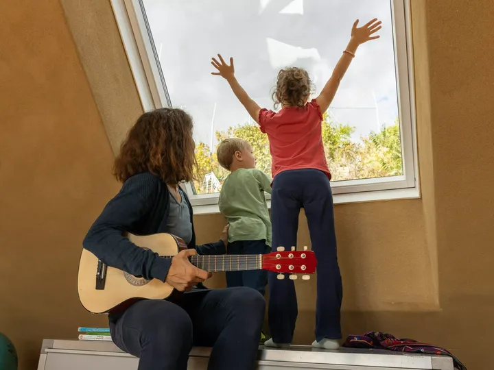 Familie genießt Musik bei einem großen Fenster, mit Kind, das die Arme dem Sonnenlicht entgegenstreckt.