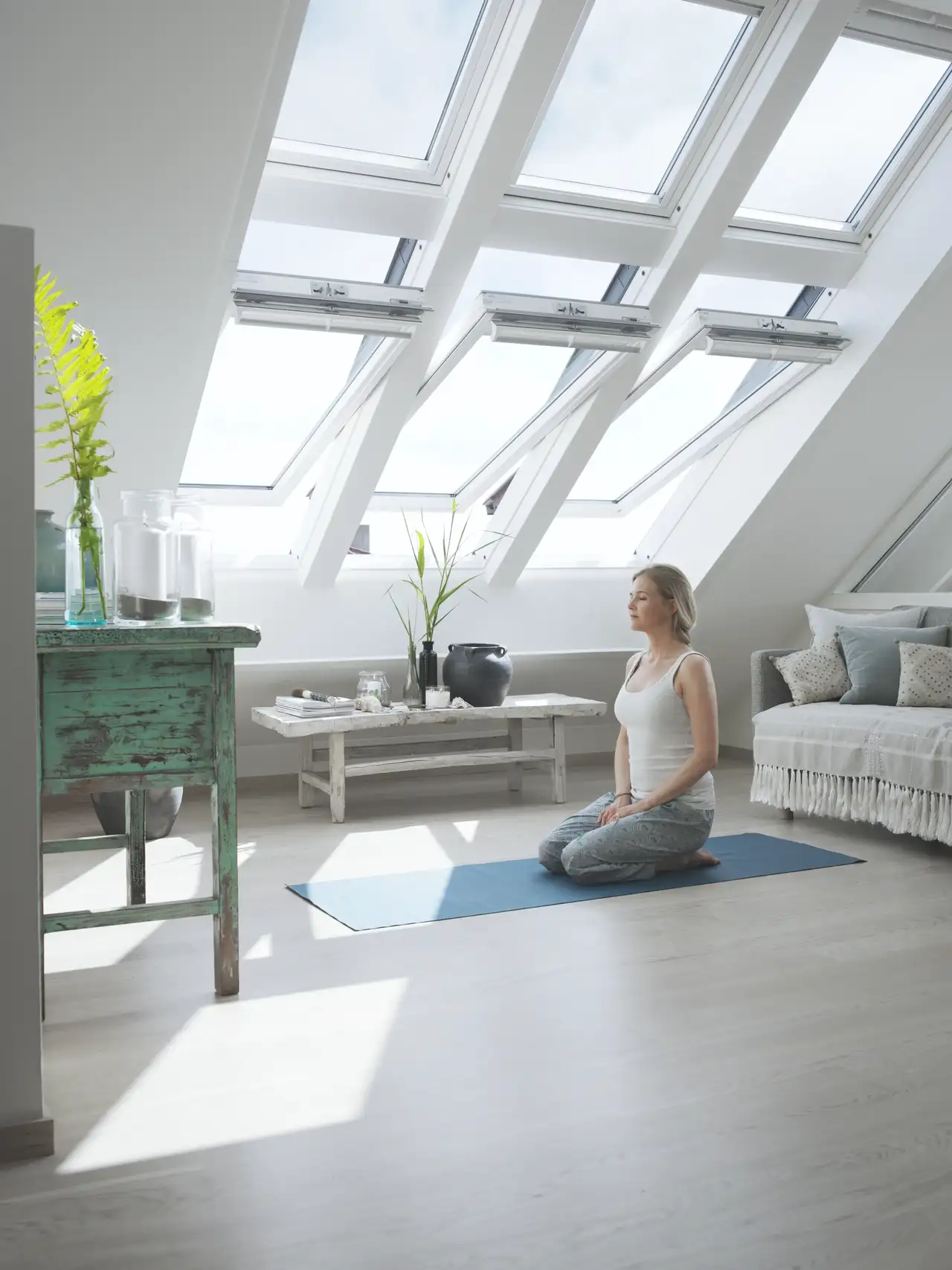 Salle lumineuse sous combles avec fenêtres de toit, mobilier minimaliste et femme en méditation sur un tapis bleu. 
