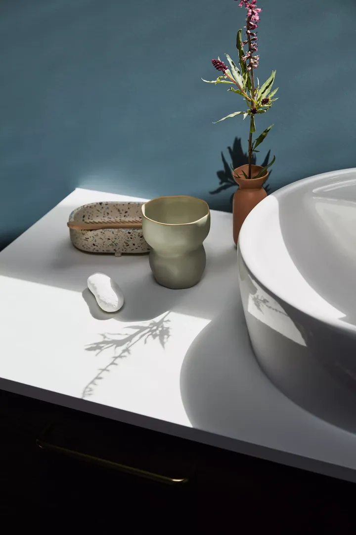 Moderner Badezimmer-Tresen mit weißem Waschbecken, grünem Becher, gesprenkeltem Tablett und Terrakotta-Vase mit Blumen.