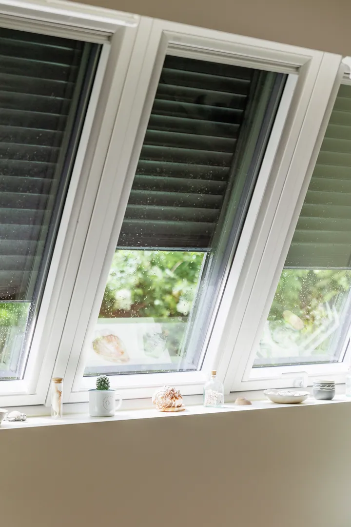 Muškarac drži daljinski upravljač dok podešava automatizovane vanjske roletne, s kapljicama kiše vidljivim na staklu. | VELUX