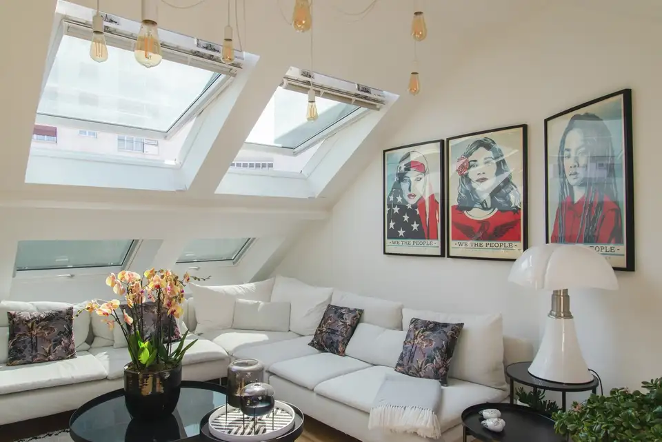 Un salon moderne sous les combles avec un canapé d'angle blanc, des fenêtres de toit VELUX, des œuvres d'art audacieuses et une lampe suspendue noire.