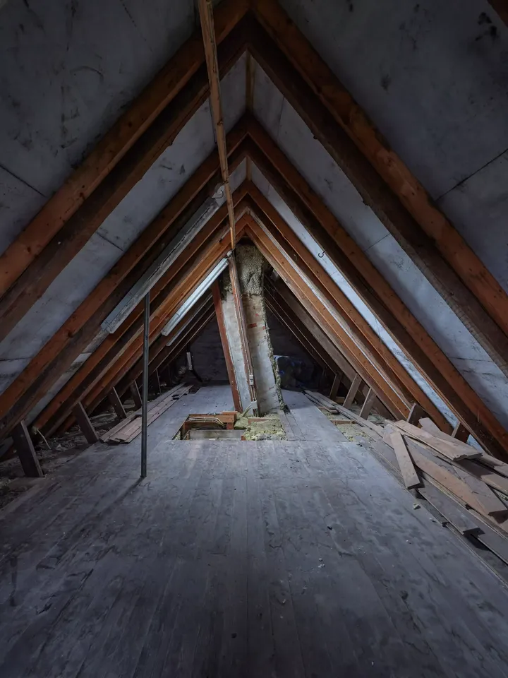 Staubiger Dachboden mit hölzernen Balken und Boden, ohne natürliches Licht.