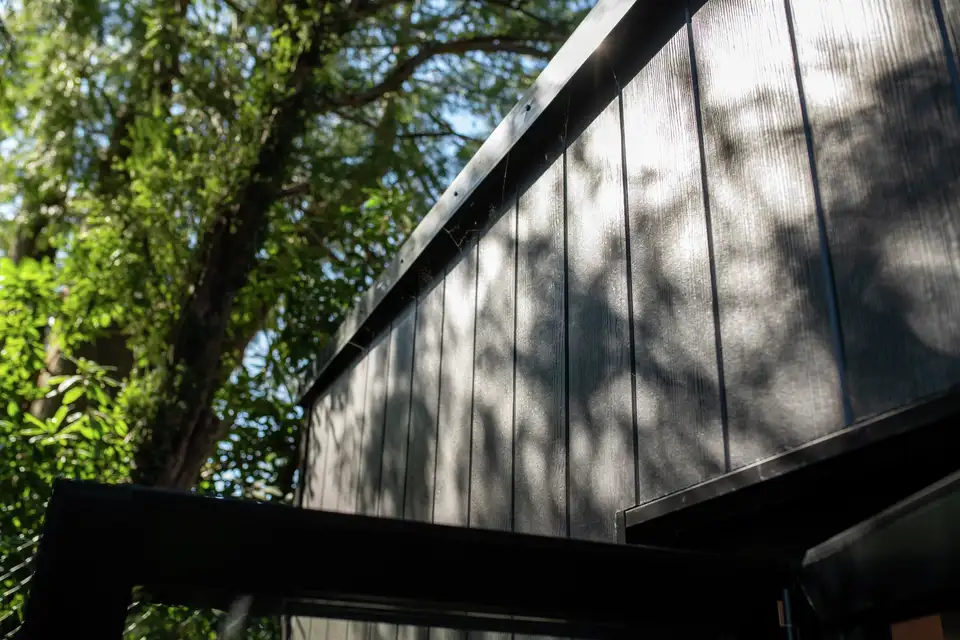 Vue en contre-plongée d’un toit de maison avec des ombres de feuilles d’arbres projetées sur sa surface, sous un ciel clair parsemé de feuillage.