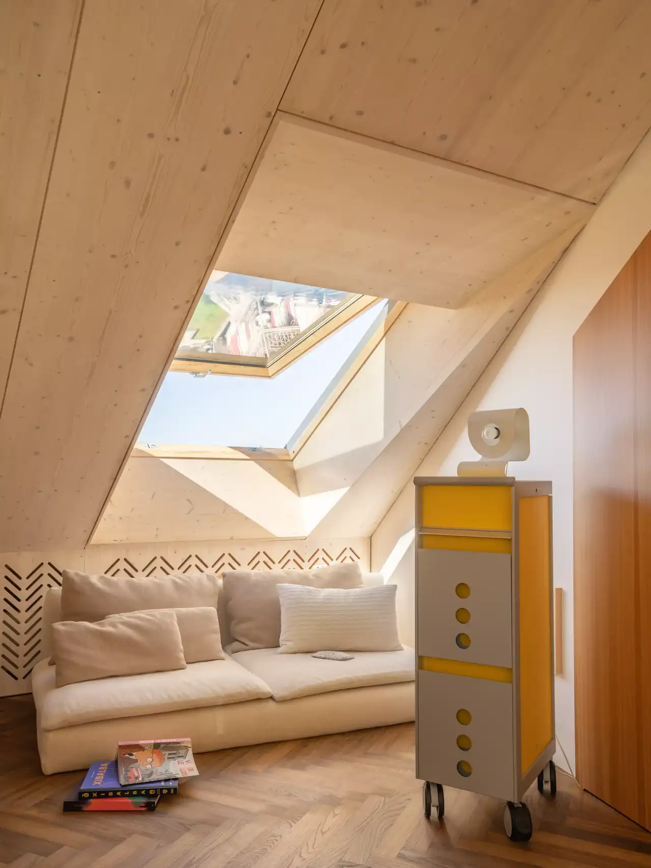 Immagine di un interno accogliente con soffitto inclinato e lucernario che lascia entrare la luce naturale. Sotto il lucernario c’è una seduta incorporata con cuscini e cassetti per lo stoccaggio. La palette dei colori è neutra con toni di legno, e lo spazio appare accogliente e ben illuminato, rendendolo una soluzione di design interessante per stanze in soffitta o a livelli superiori.