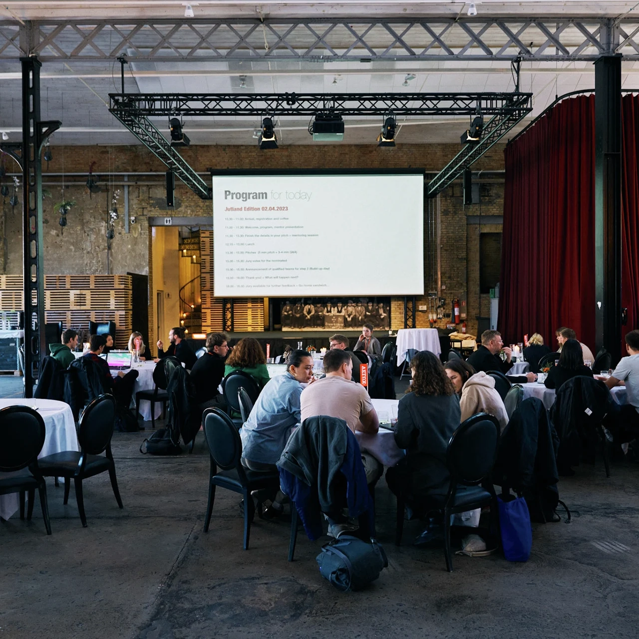 Conference attendees seated in an industrial hall with a large screen.