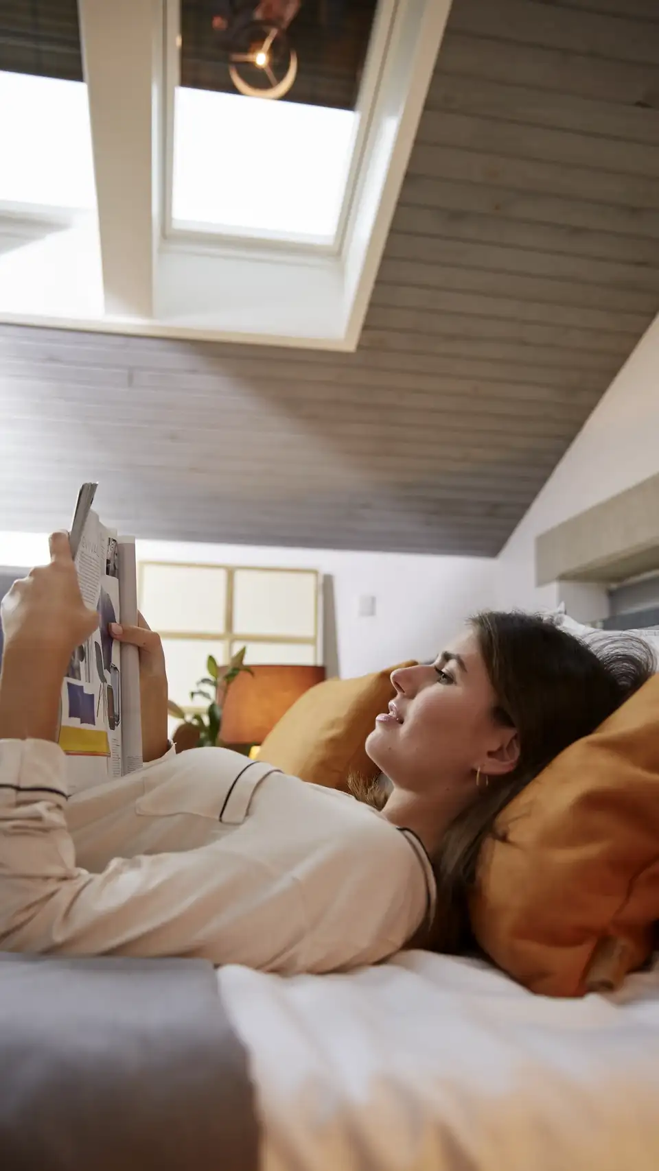  une femme lit un magazine au lit dans une chambre équipée de fenêtres de toit VELUX