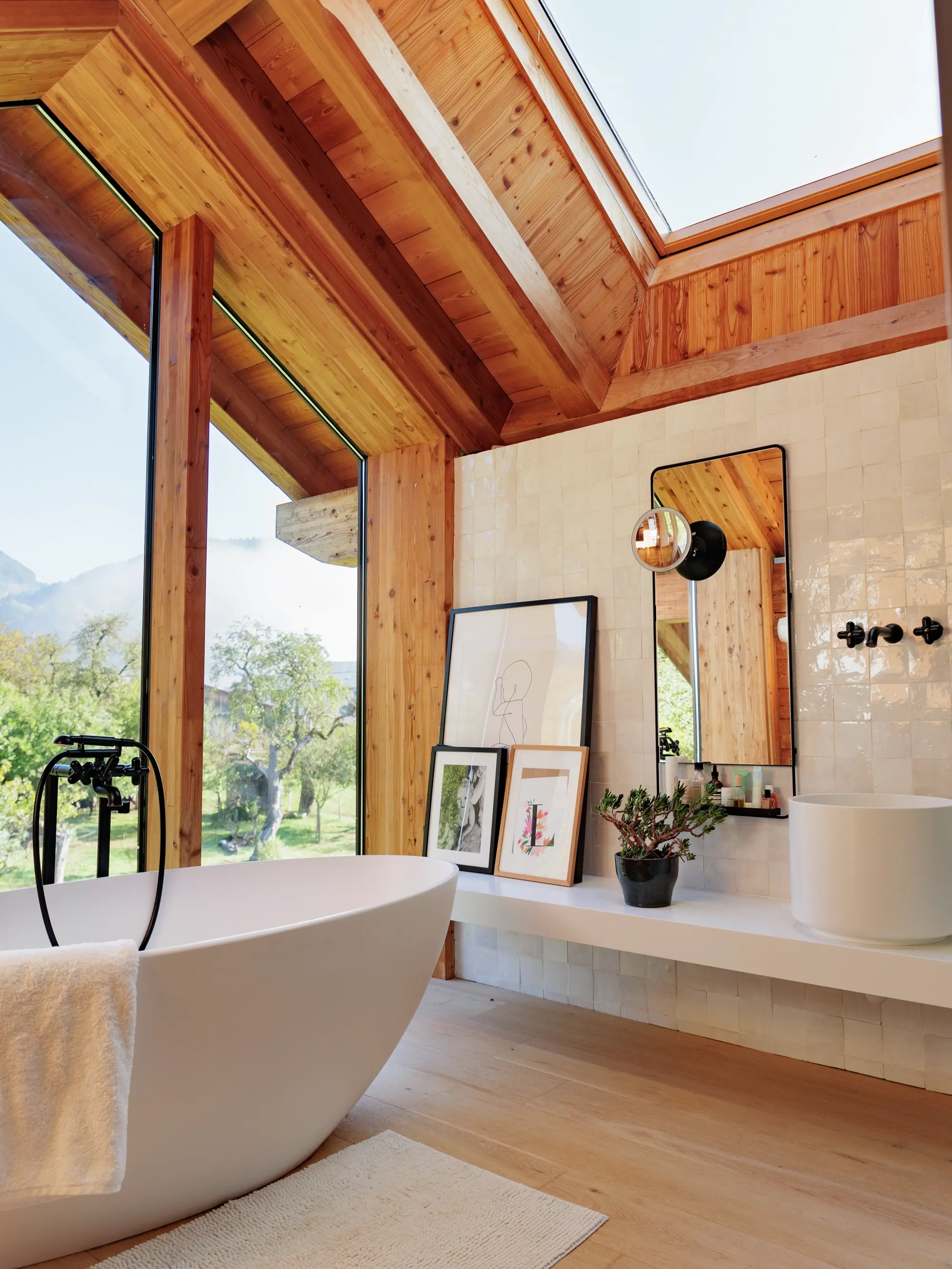 Salle de bain moderne avec une baignoire indépendante, des murs carrelés en beige, un sol en bois et un plafond en bois avec de grandes fenêtres donnant sur un paysage naturel.