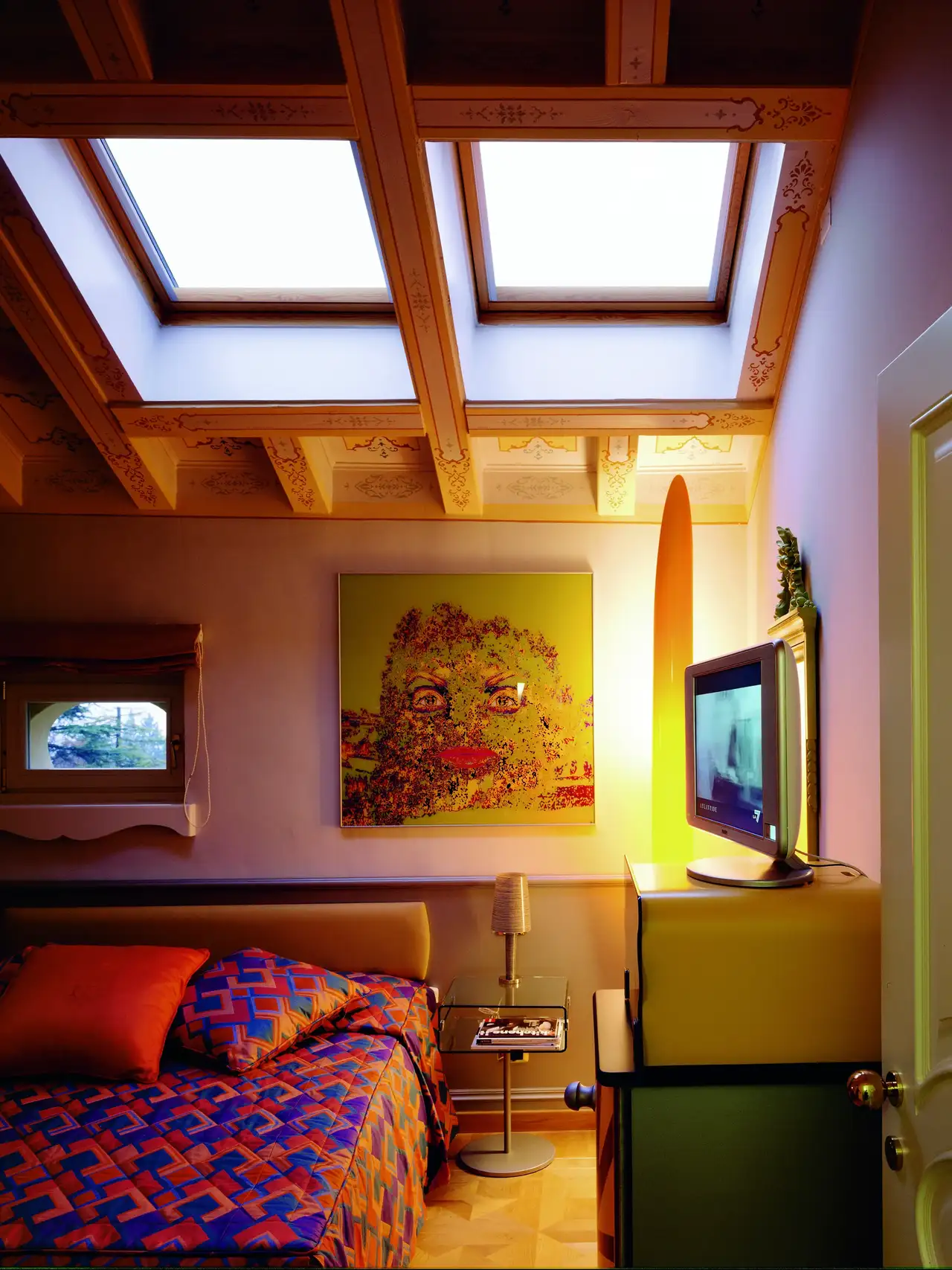 Chambre mansardée avec couvre-lit géométrique coloré, meubles vintage et lumière naturelle des fenêtres de toit.