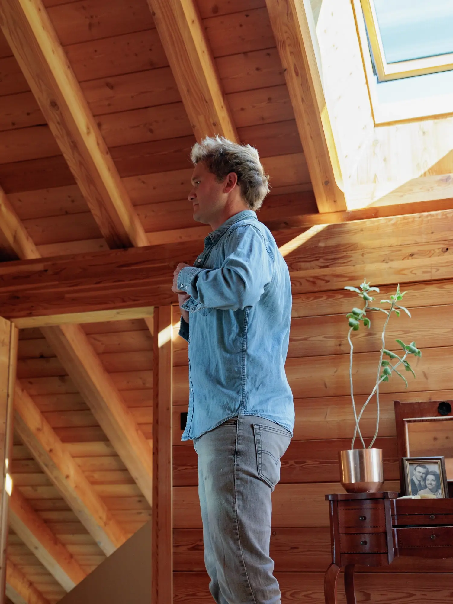 Jean Philippe debout dans un intérieur chaleureux avec des murs et un plafond en bois, près d'une fenêtre de toit qui offre une isolation efficace, ainsi qu'une abondance de lumière naturelle et d'air frais.