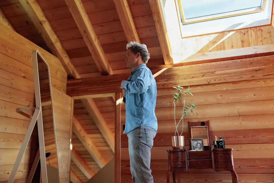 Uomo si abbottona la camicia azzurra in camera sotto una finestra per tetti VELUX.