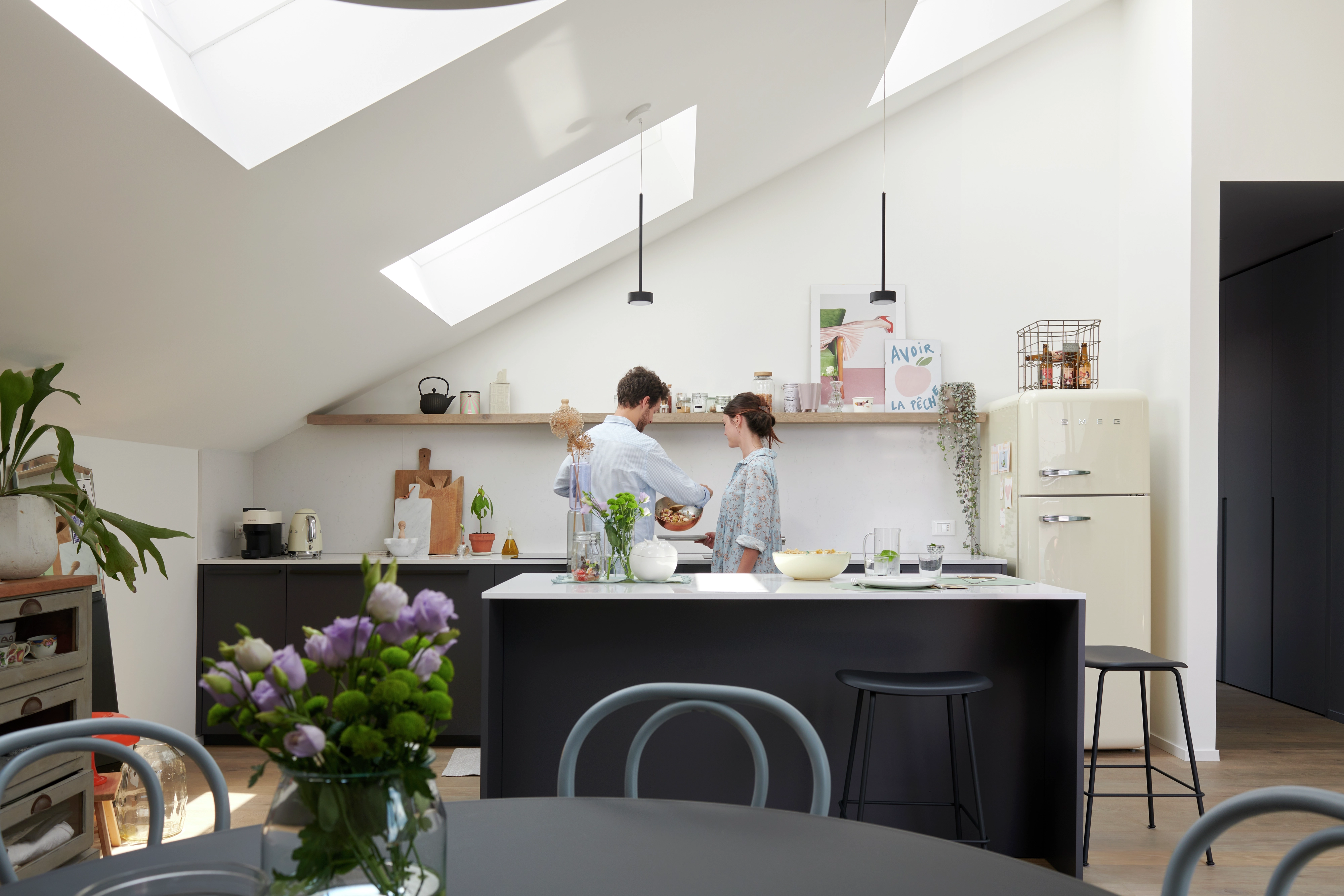 Cucina moderna e luminosa con finestre per tetti VELUX, piani di lavoro neri e frigorifero color crema.